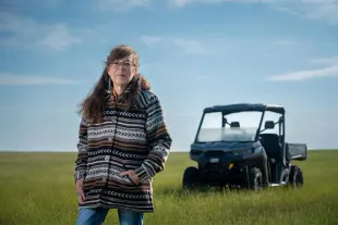 Person wearing glasses and patterned cardigan standing in a vast green field with a utility vehicle in the background under a blue sky.
