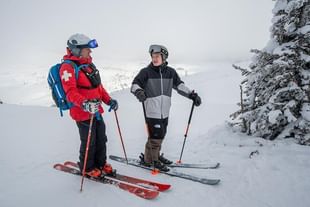 Two skiers stand on a snowy mountain slope wearing winter gear and helmets, with snow-covered evergreen trees visible on the right and a scenic valley landscape in the background.