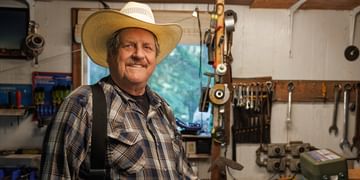Man wearing a cowboy hat and plaid shirt smiling in a workshop filled with tools, equipment, and hanging instruments on the walls