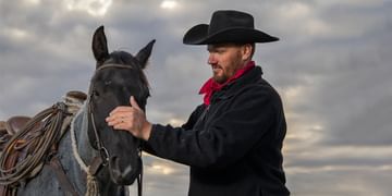 STARS Very Important Patient and rancher Milt Mastad strokes the nose of his saddled grey horse in front of a cloudy sky.