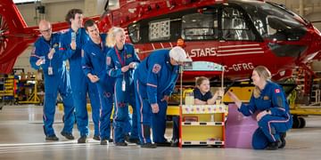 STARS operations staff line up to buy lemonade from STARS Very Important Patient Alexis Moore's lemonade stand setup in a STARS hangar in front of a STARS air ambulance helicopter.