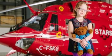 STARS Very Important Patient Breanna Booy holds her Starbear teddy that she got during her flight in a STARS air ambulance. She smiles for the camera while posing in front of a STARS BK117 air ambulance helicopter in this older photo.
