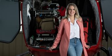 STARS Very Important Patient Carla Minogue leans against the back of a STARS air ambulance helicopter. The rear clamshell doors are open, and the entire medical cabin is visible behind her.