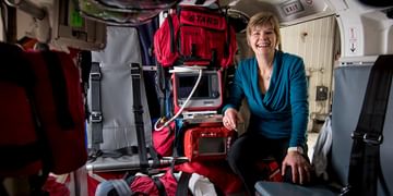 STARS Very Important Patient Charlotte McHugh sits inside the medical cabin of a STARS air ambulance helicopter, laughing at something said by someone off screen. Various pieces of medical equipment are visible in the cabin.