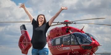 STARS Very Important Patient Chelsie Bullock jumps in the air with her hands up in front of a STARS air ambulance helicopter on a cloudy day.