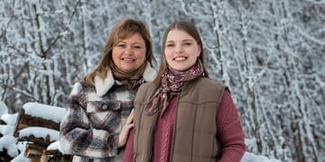 Esther and Kaycee Stern pose together during a cool winter day at their property. Snow covered trees and logs are in the background.