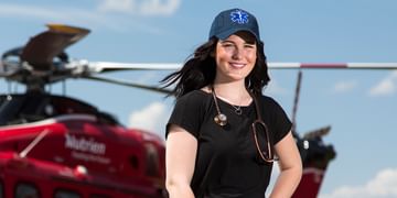STARS Very Important Patient Jessi Fredin wears an EMS ballcap and a stethoscope while standing in front of an older STARS AW139 air ambulance helicopter on a blue sky day.