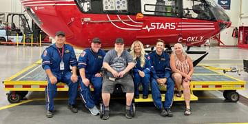 STARS Very Important Patient Tyler Lister sits together with STARS crew that flew on his mission, and his mother, in front of a red and black helicopter labeled STARS in an aircraft hangar.