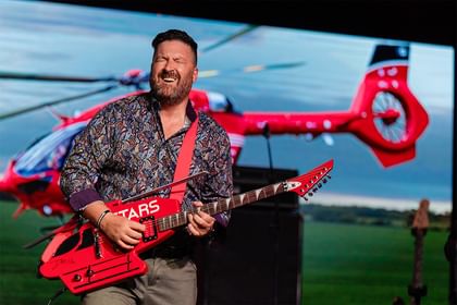 A guitarist plays a custom made guitar made to resemble a red STARS helicopter on stage at a past STARS event.