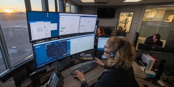 STARS Vigilant emergency communications specialists work at double curved monitor workstations at sunrise, looking over the airfield from the Vigilant Emergency Communications Centre. On the screens are the various windows that staff use to monitor sites, emergency response plans, and workers alone in the field.