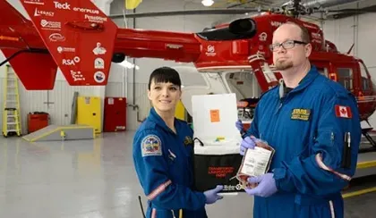 Two STARS crew members in blue flight suits pose with clinically packaged human blood in front of a red helicopter inside a hangar.