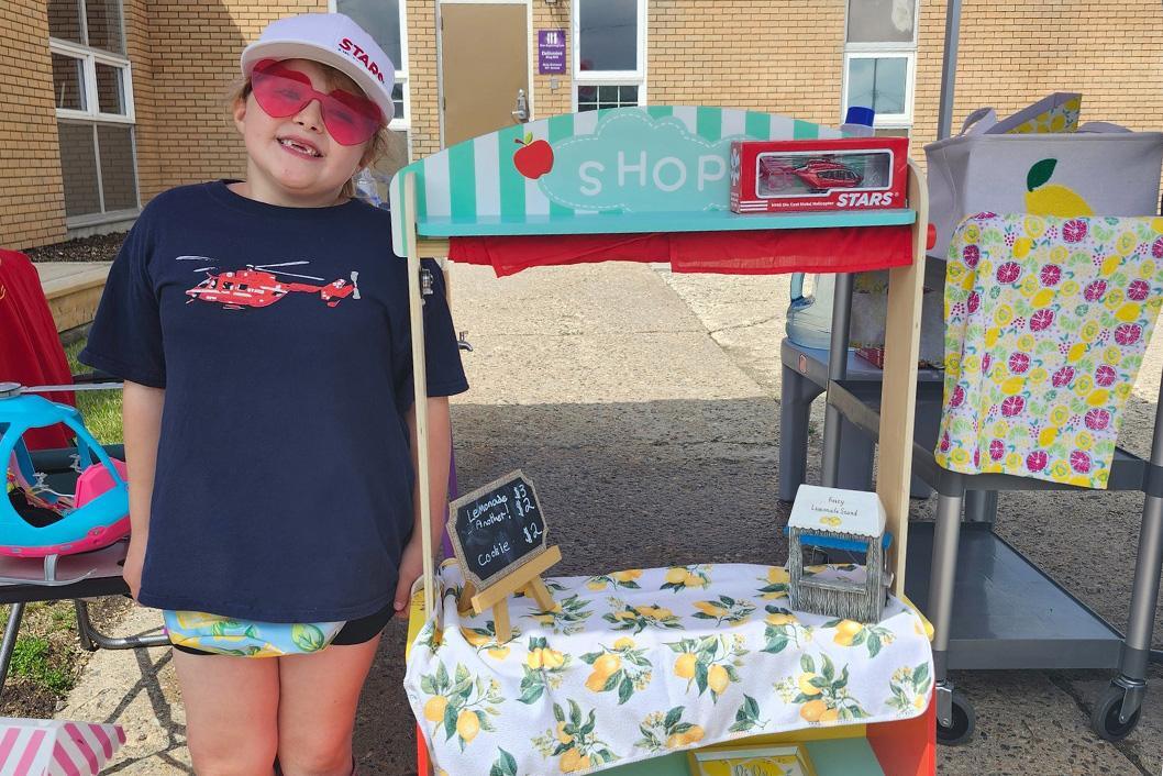 STARS Very Important Patient and Ally Alexis Moore stands next to her Lemonade stand she's called Sips for STARS. She wears a STARS helicopter tee while a boxed STARS H145 air ambulance helicopter toy sits on top of the stand.