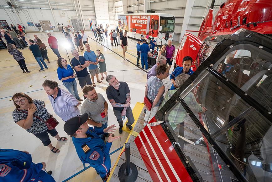 Attendees in the foreground look inside a STARS H145 air ambulance helicopter while a member of the STARS operations team details working inside. The photo is taken from an elevated angle, in the background other attendees look at exhibits and table displays setup during the Open House event held at the Calgary base in celebration of STARS' 40th anniversary.